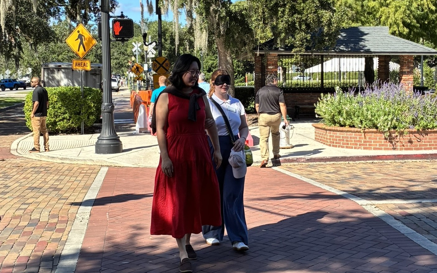 A blindfolded woman is guided across the street by a woman in a red dress.